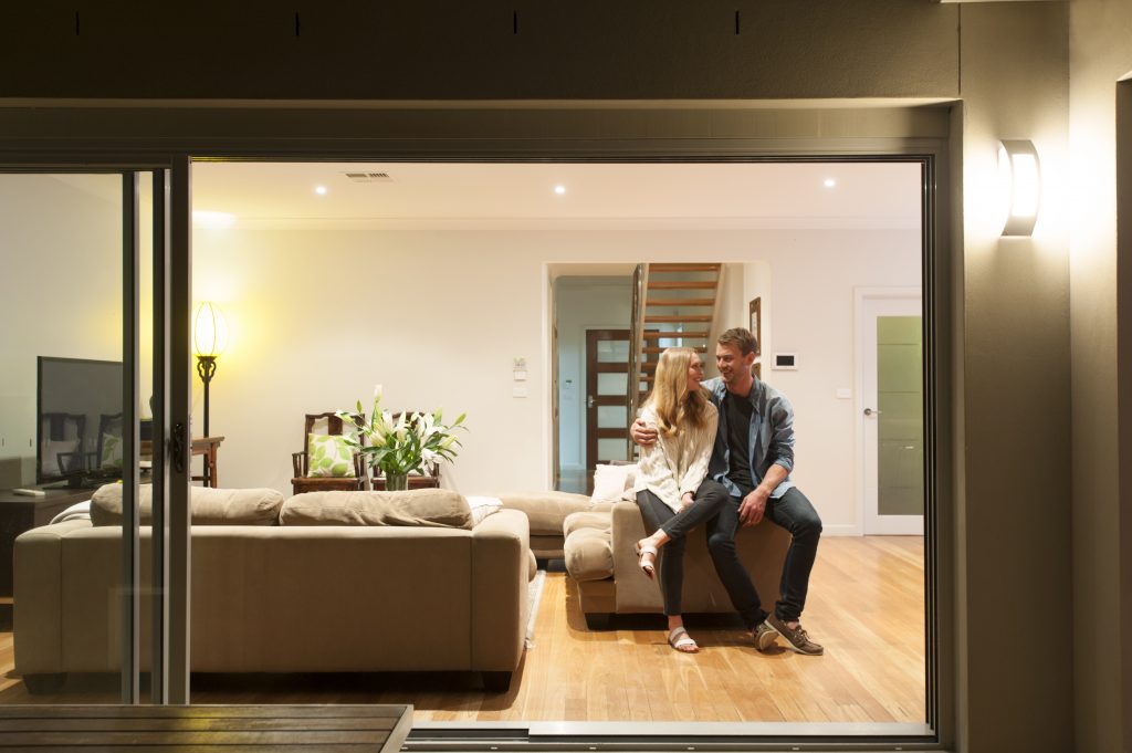 Couple relaxing in their home at night. They are both wearing casual clothes and embracing. They are looking at each other and smiling. The house is contemporary with an open plan al fresco style. Copy space